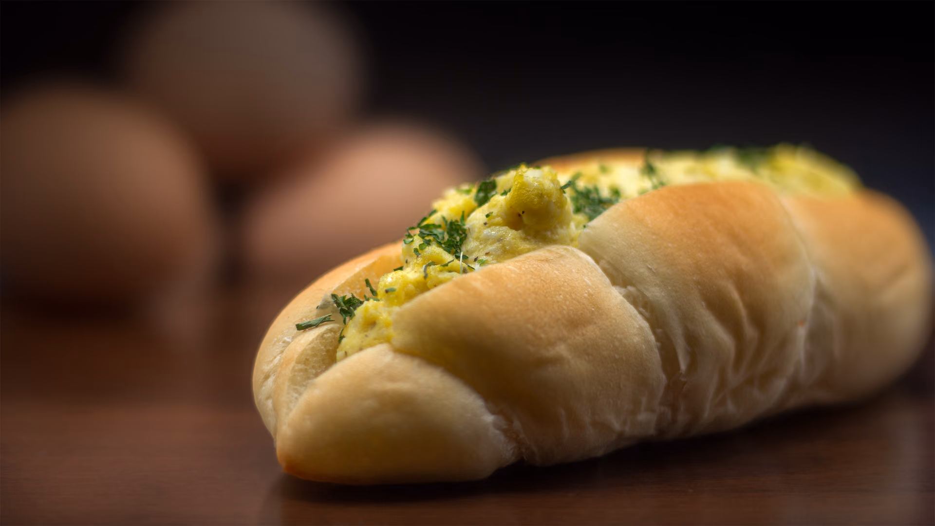 A soft split roll filled with herb-flecked egg salad, whole eggs blurred behind it on dark walnut.